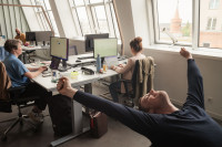 Leute sitzen im Büro und arbeiten vor ihren Bildschirmen. Im Vordergrund streckt sich ein Mitarbeiter und lächelt.